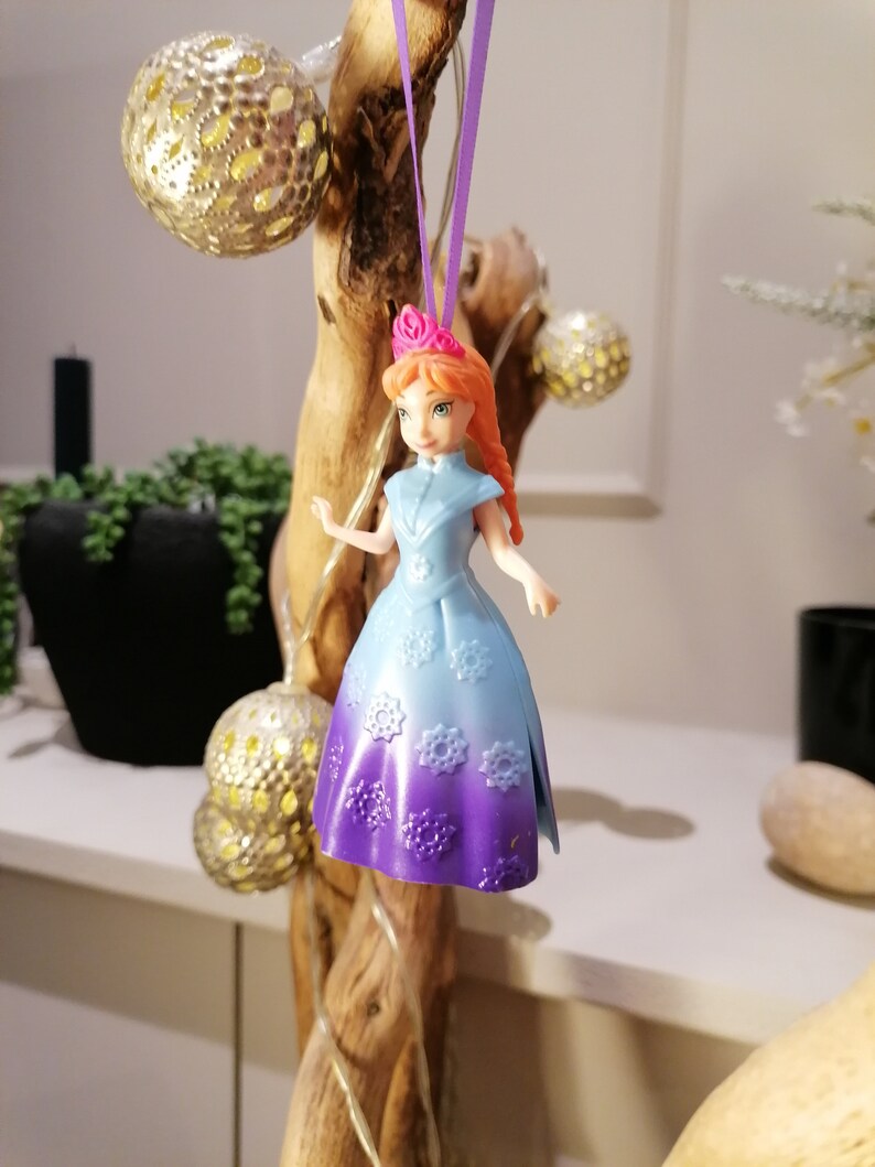 K&ouml;nnte beinhalten: Eine Plastikpuppe mit roten Haaren und einer rosa Krone, die ein lila-blaues Kleid mit Schneeflockenmustern tr&auml;gt. Die Puppe h&auml;ngt an einem lila Band.