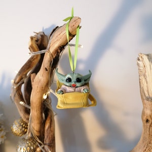 May include: A green and brown Christmas ornament featuring a character from the Star Wars franchise, sitting in a yellow bag. The ornament is hanging from a green ribbon on a branch.