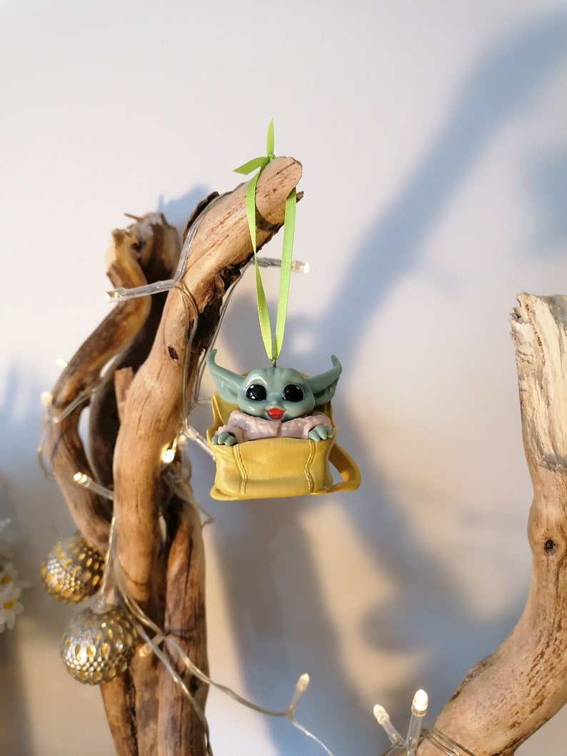 May include: A green and brown Christmas ornament featuring a character from the Star Wars franchise, sitting in a yellow bag. The ornament is hanging from a green ribbon on a branch.