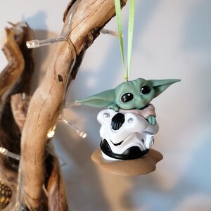 K&ouml;nnte beinhalten: Ein gr&uuml;ner und brauner Weihnachtsbaumschmuck mit einer Figur aus dem Star Wars-Franchise, The Mandalorian. Die Figur sitzt in einem kleinen, wei&szlig;en, podartigen Fahrzeug.