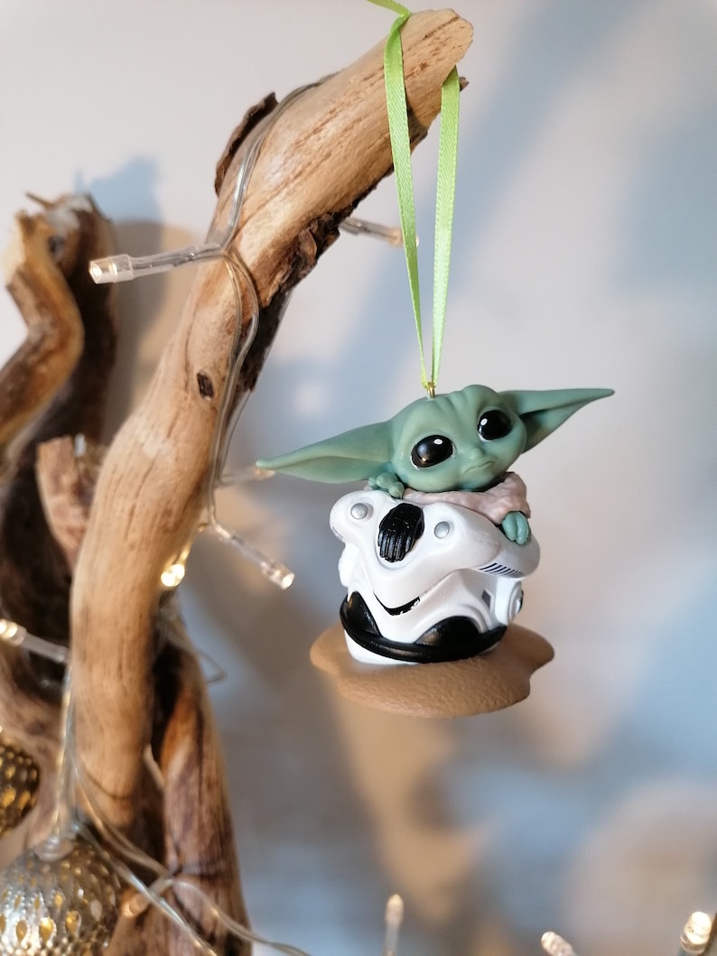 K&ouml;nnte beinhalten: Ein gr&uuml;ner und brauner Weihnachtsbaumschmuck mit einer Figur aus dem Star Wars-Franchise, The Mandalorian. Die Figur sitzt in einem kleinen, wei&szlig;en, podartigen Fahrzeug.