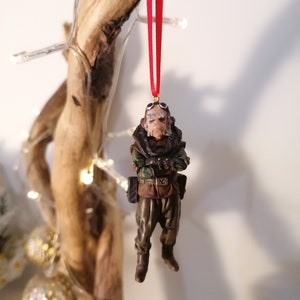 K&ouml;nnte beinhalten: Detaillierte Figur einer Figur mit Brille und milit&auml;rischer Kleidung, die an einem roten Band h&auml;ngt. Die Figur ist braun, gr&uuml;n und beige, mit einem strengen Ausdruck. Der Schmuck ist wahrscheinlich f&uuml;r einen Weihnachtsbaum.