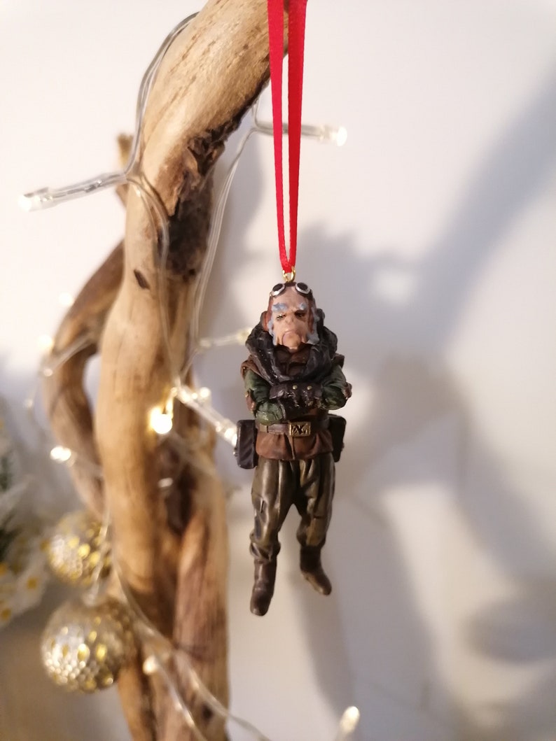 K&ouml;nnte beinhalten: Detaillierte Figur einer Figur mit Brille und milit&auml;rischer Kleidung, die an einem roten Band h&auml;ngt. Die Figur ist braun, gr&uuml;n und beige, mit einem strengen Ausdruck. Der Schmuck ist wahrscheinlich f&uuml;r einen Weihnachtsbaum.