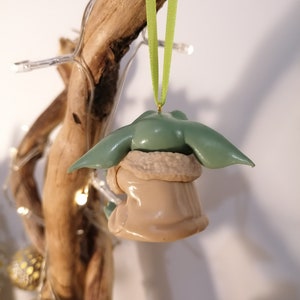 May include: A green and brown Christmas ornament shaped like a character from the Star Wars franchise. The ornament is hanging from a green ribbon.