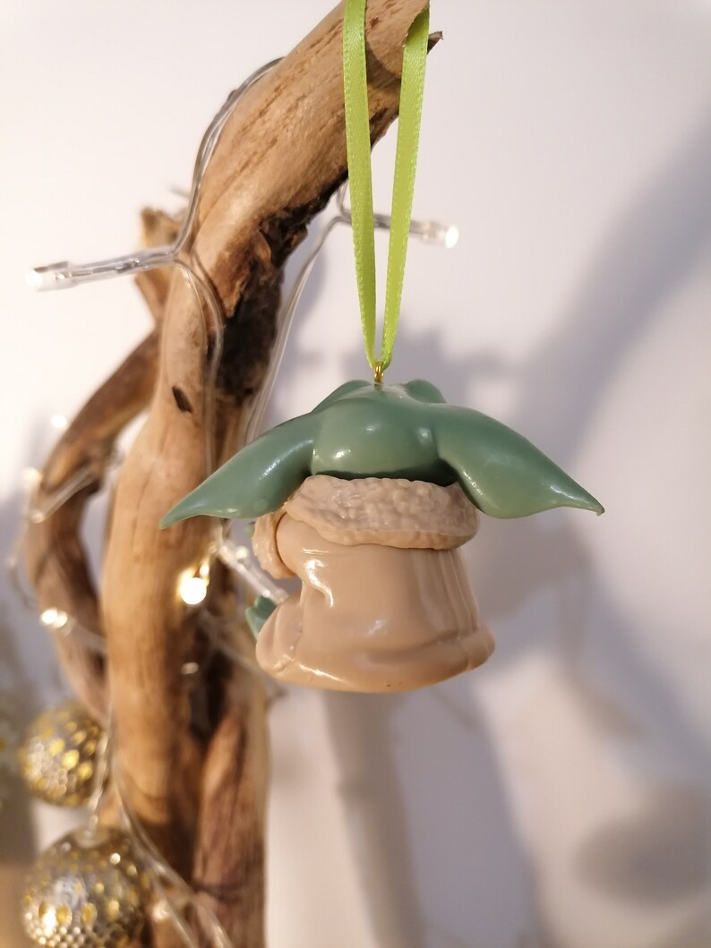 May include: A green and brown Christmas ornament shaped like a character from the Star Wars franchise. The ornament is hanging from a green ribbon.