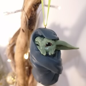 May include: A green and blue Christmas ornament featuring a small, green alien-like character wrapped in a blue blanket. The ornament is hanging from a green ribbon.