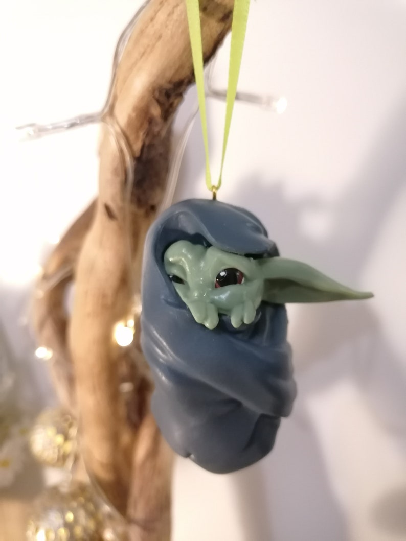 May include: A green and blue Christmas ornament featuring a small, green alien-like character wrapped in a blue blanket. The ornament is hanging from a green ribbon.