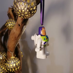 May include: A white and green plastic toy astronaut ornament hanging from a purple ribbon. The ornament is a character from the Toy Story movie franchise.