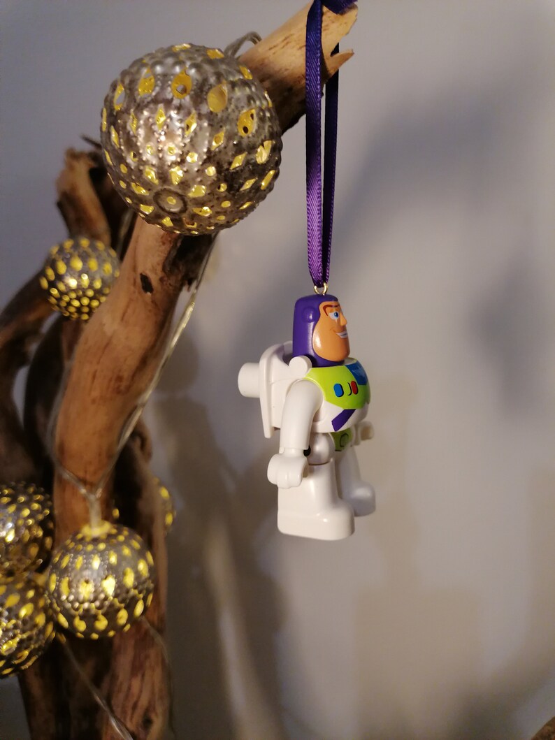 May include: A white and green plastic toy astronaut ornament hanging from a purple ribbon. The ornament is a character from the Toy Story movie franchise.