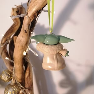 May include: A green and beige Christmas ornament shaped like a character from the Star Wars franchise. The ornament is hanging from a green ribbon on a branch.