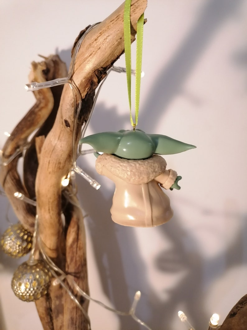 May include: A green and beige Christmas ornament shaped like a character from the Star Wars franchise. The ornament is hanging from a green ribbon on a branch.