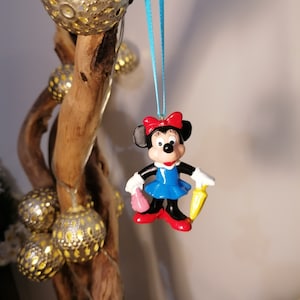 Op de afbeelding: Een blauwe en rode plastic figuur van Minnie Mouse die een gele paraplu en een roze handtas vasthoudt. De figuur hangt aan een blauw lint.