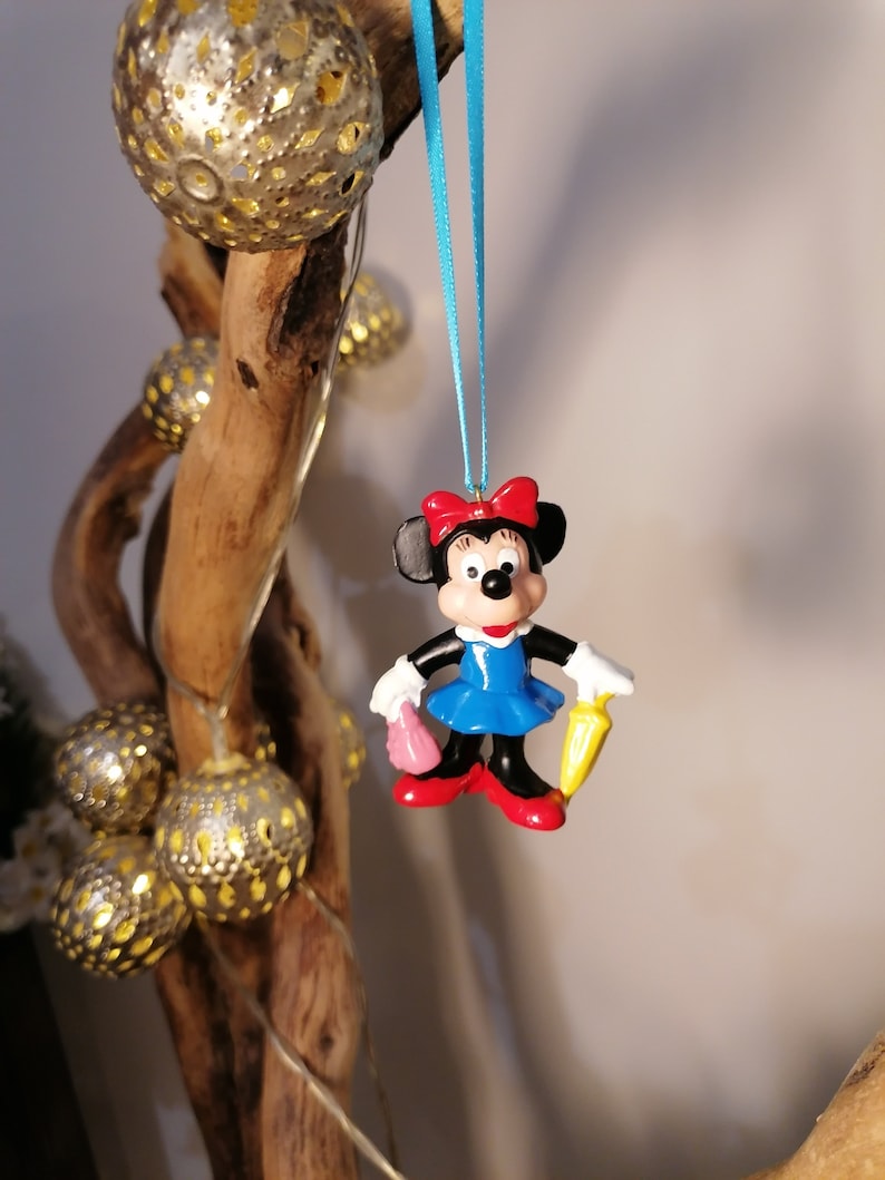 Op de afbeelding: Een blauwe en rode plastic figuur van Minnie Mouse die een gele paraplu en een roze handtas vasthoudt. De figuur hangt aan een blauw lint.