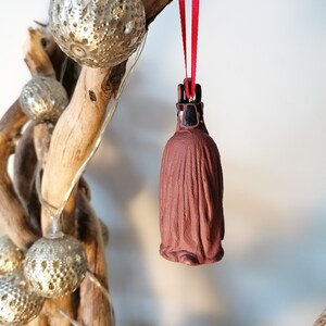 May include: A brown ceramic ornament shaped like a hooded figure with a red ribbon hanging from the top. The ornament is hanging from a branch with silver and white decorative lights.