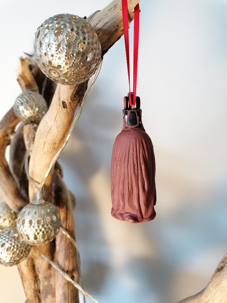May include: A brown ceramic ornament shaped like a hooded figure with a red ribbon hanging from the top. The ornament is hanging from a branch with silver and white decorative lights.