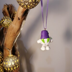 May include: A purple and white toy astronaut ornament with a green visor hanging from a purple ribbon. The ornament is hanging from a branch with gold-colored ornaments.