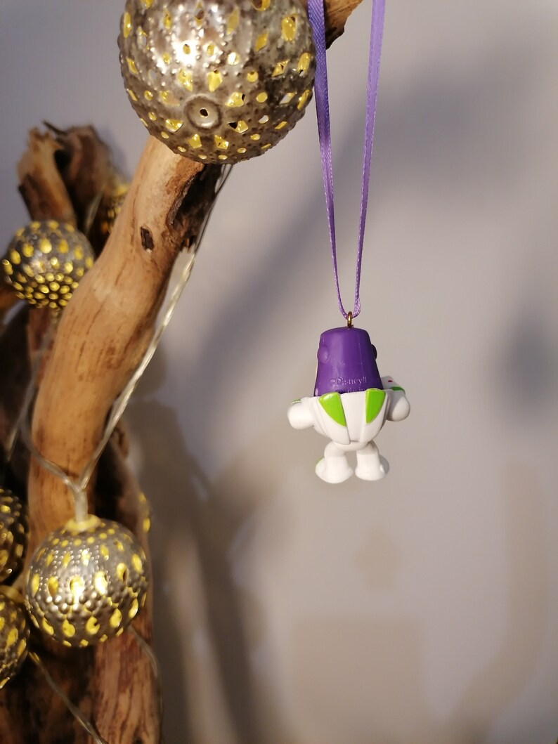 May include: A purple and white toy astronaut ornament with a green visor hanging from a purple ribbon. The ornament is hanging from a branch with gold-colored ornaments.