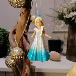 Peut inclure: Une figurine de poup&eacute;e v&ecirc;tue d'une robe blanche et bleue avec une couronne bleue. La poup&eacute;e a les cheveux blonds, les yeux bleus et une robe blanche avec une jupe bleu d&eacute;grad&eacute;.