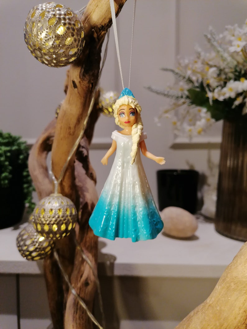Peut inclure: Une figurine de poup&eacute;e v&ecirc;tue d'une robe blanche et bleue avec une couronne bleue. La poup&eacute;e a les cheveux blonds, les yeux bleus et une robe blanche avec une jupe bleu d&eacute;grad&eacute;.
