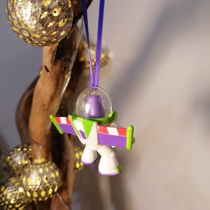 Puede incluir: Un adorno de pl&aacute;stico morado, verde y blanco de Buzz Lightyear de la franquicia Toy Story. El adorno est&aacute; colgado de una cinta morada.