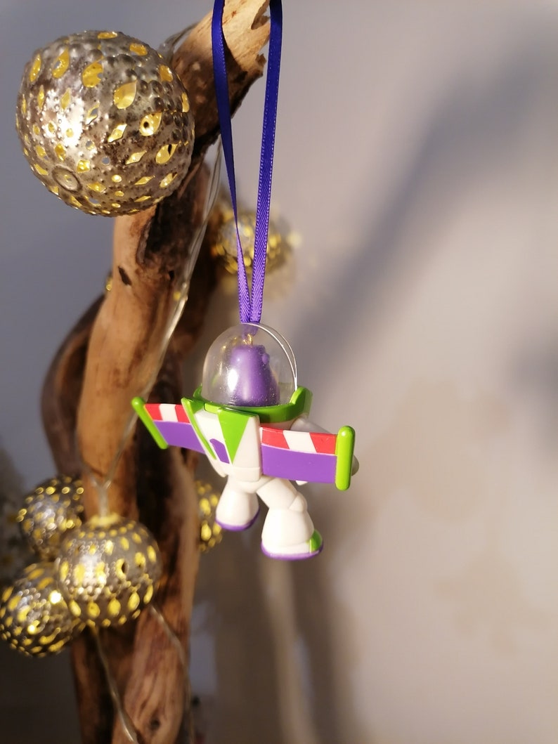 Puede incluir: Un adorno de pl&aacute;stico morado, verde y blanco de Buzz Lightyear de la franquicia Toy Story. El adorno est&aacute; colgado de una cinta morada.