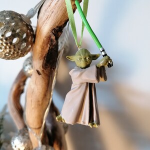 Puede incluir: Adorno navide&ntilde;o de Yoda con un sable de luz verde, colgado de una cinta verde claro. La figura de Yoda es verde y marr&oacute;n, con una t&uacute;nica marr&oacute;n. El adorno cuelga de un trozo de madera flotante.