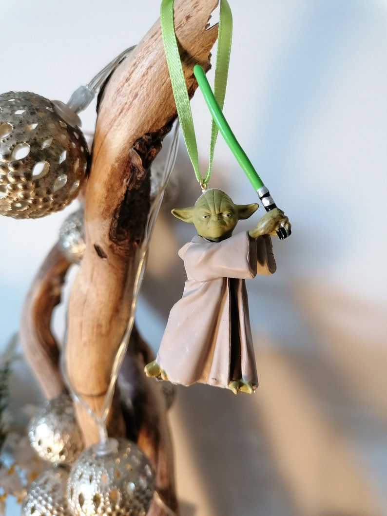 Puede incluir: Adorno navide&ntilde;o de Yoda con un sable de luz verde, colgado de una cinta verde claro. La figura de Yoda es verde y marr&oacute;n, con una t&uacute;nica marr&oacute;n. El adorno cuelga de un trozo de madera flotante.