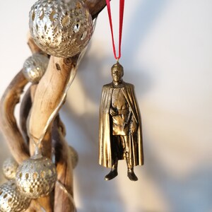 May include: A gold-colored ornament depicting a person wearing a long robe and a crown. The ornament is hanging from a red ribbon.