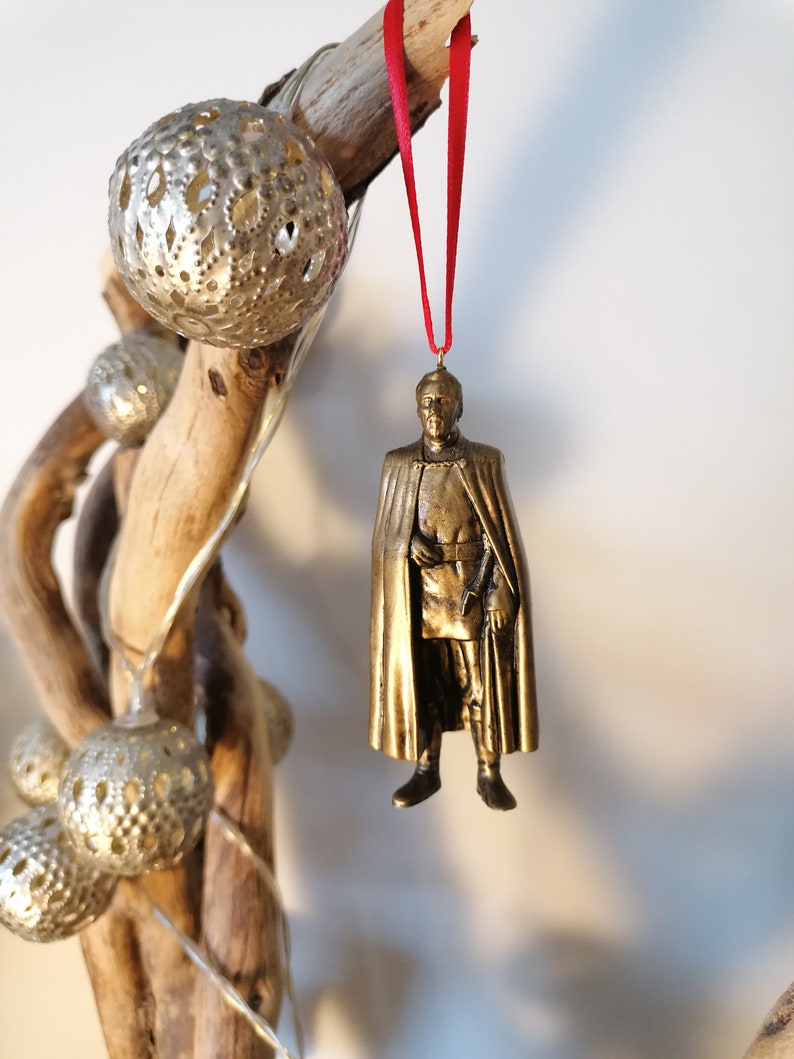 May include: A gold-colored ornament depicting a person wearing a long robe and a crown. The ornament is hanging from a red ribbon.