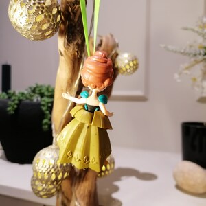 May include: A small plastic doll of a princess with brown hair, wearing a teal and gold dress, hanging from a green ribbon.