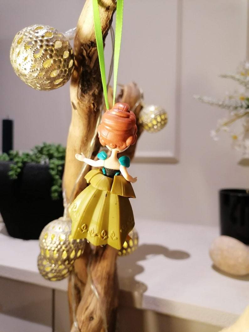 May include: A small plastic doll of a princess with brown hair, wearing a teal and gold dress, hanging from a green ribbon.