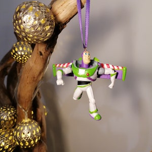May include: A purple and green plastic toy figurine of Buzz Lightyear from the Toy Story movie franchise. The figurine is hanging from a purple ribbon and has wings.