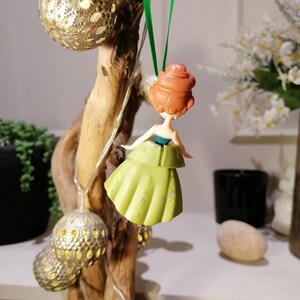 May include: A green and gold Christmas ornament featuring a cartoon princess with red hair. The ornament is hanging from a green string.