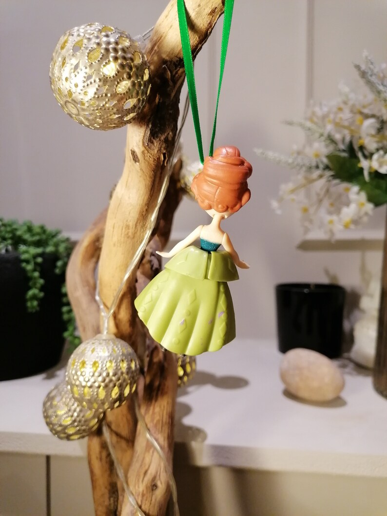 May include: A green and gold Christmas ornament featuring a cartoon princess with red hair. The ornament is hanging from a green string.