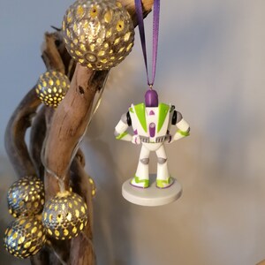 May include: A white and green Buzz Lightyear Christmas ornament with purple accents. The ornament is hanging from a purple ribbon on a branch with decorative lights.