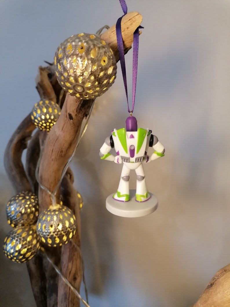 May include: A white and green Buzz Lightyear Christmas ornament with purple accents. The ornament is hanging from a purple ribbon on a branch with decorative lights.