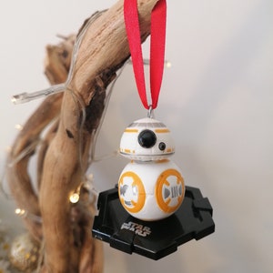 May include: A white and orange Star Wars BB-8 droid Christmas ornament with a black hexagonal base. The ornament is hanging from a red ribbon.