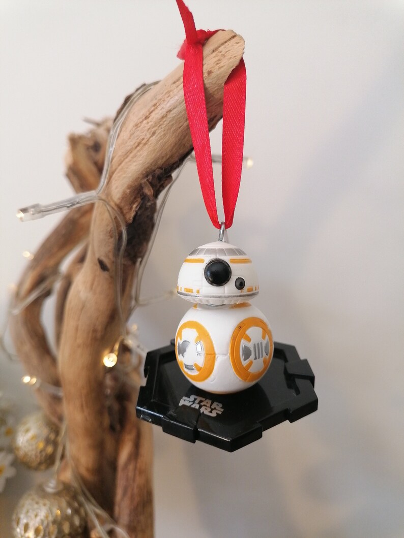 May include: A white and orange Star Wars BB-8 droid Christmas ornament with a black hexagonal base. The ornament is hanging from a red ribbon.