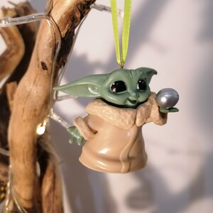 May include: A green and brown Christmas ornament featuring a character from the Star Wars franchise, holding a silver ball.