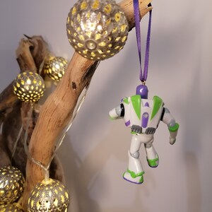 May include: A white and green Buzz Lightyear toy ornament with purple accents hanging from a purple ribbon on a branch with fairy lights and silver baubles.