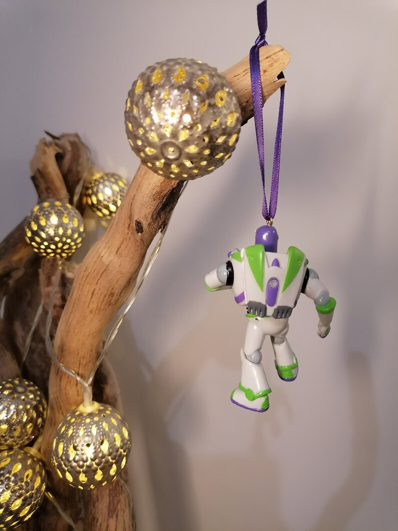 May include: A white and green Buzz Lightyear toy ornament with purple accents hanging from a purple ribbon on a branch with fairy lights and silver baubles.
