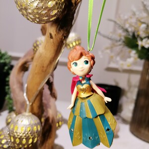 Pu&ograve; includere: Un ornamento a corda verde con una piccola figurina di plastica di una principessa che indossa un abito verde acqua e oro con un mantello rosso. La principessa ha i capelli castani, gli occhi azzurri e un leggero blush rosa.