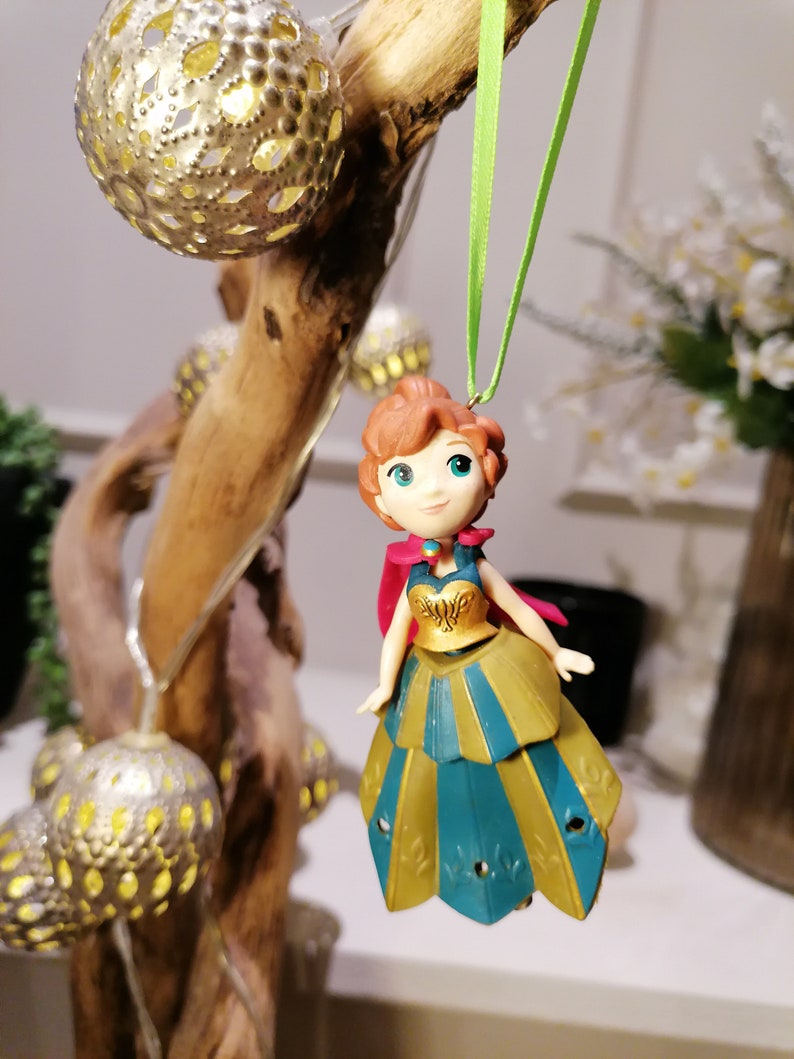 Pu&ograve; includere: Un ornamento a corda verde con una piccola figurina di plastica di una principessa che indossa un abito verde acqua e oro con un mantello rosso. La principessa ha i capelli castani, gli occhi azzurri e un leggero blush rosa.