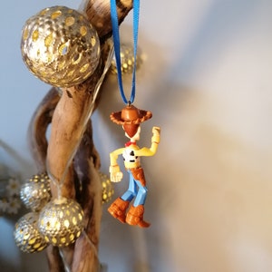 May include: A blue ribbon hangs from a piece of driftwood with a small plastic figurine of Woody from Toy Story attached. Woody is wearing his signature cowboy hat, vest, and jeans.