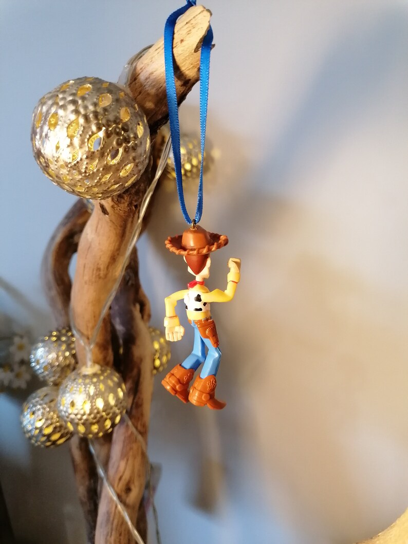 May include: A blue ribbon hangs from a piece of driftwood with a small plastic figurine of Woody from Toy Story attached. Woody is wearing his signature cowboy hat, vest, and jeans.