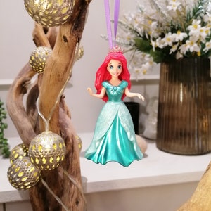 May include: A red-haired Disney princess doll wearing a turquoise gown and a crown. The doll is hanging from a purple ribbon.