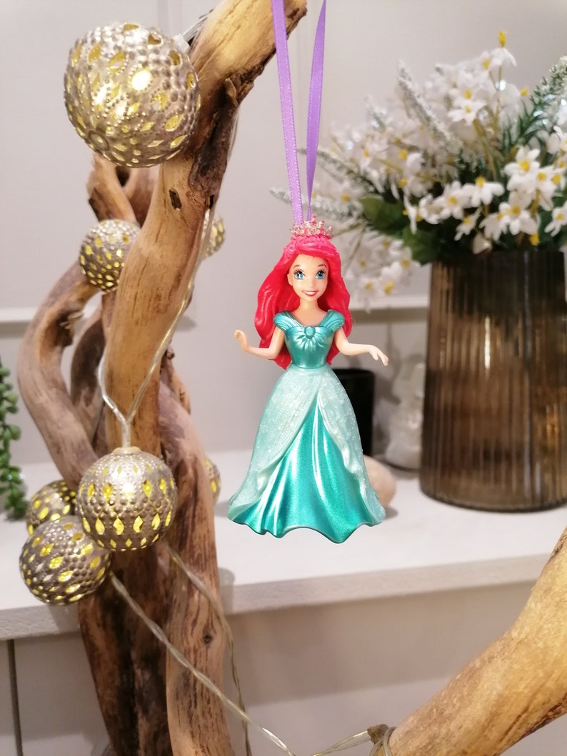 May include: A red-haired Disney princess doll wearing a turquoise gown and a crown. The doll is hanging from a purple ribbon.