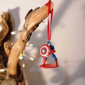 May include: A red, white, and blue Captain America Christmas ornament with a red ribbon hanging from a branch.