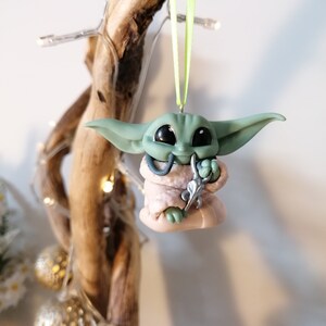 May include: A green and pink Christmas ornament shaped like a character from the Star Wars franchise. The ornament is hanging from a green ribbon.