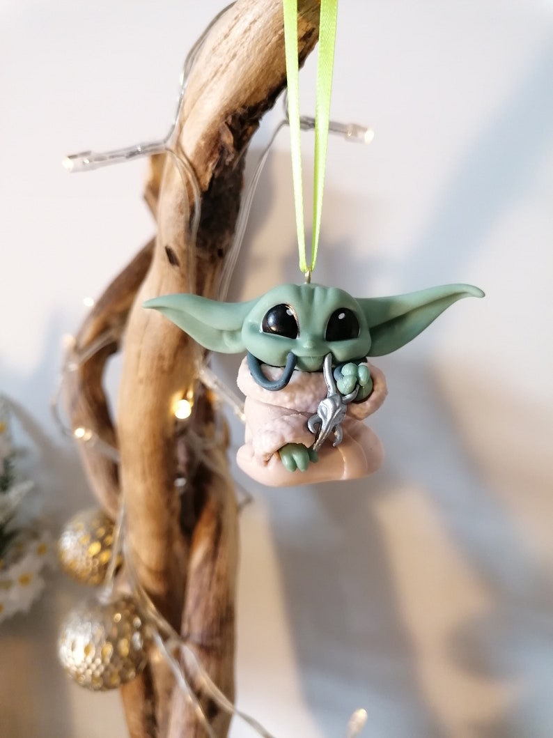 May include: A green and pink Christmas ornament shaped like a character from the Star Wars franchise. The ornament is hanging from a green ribbon.